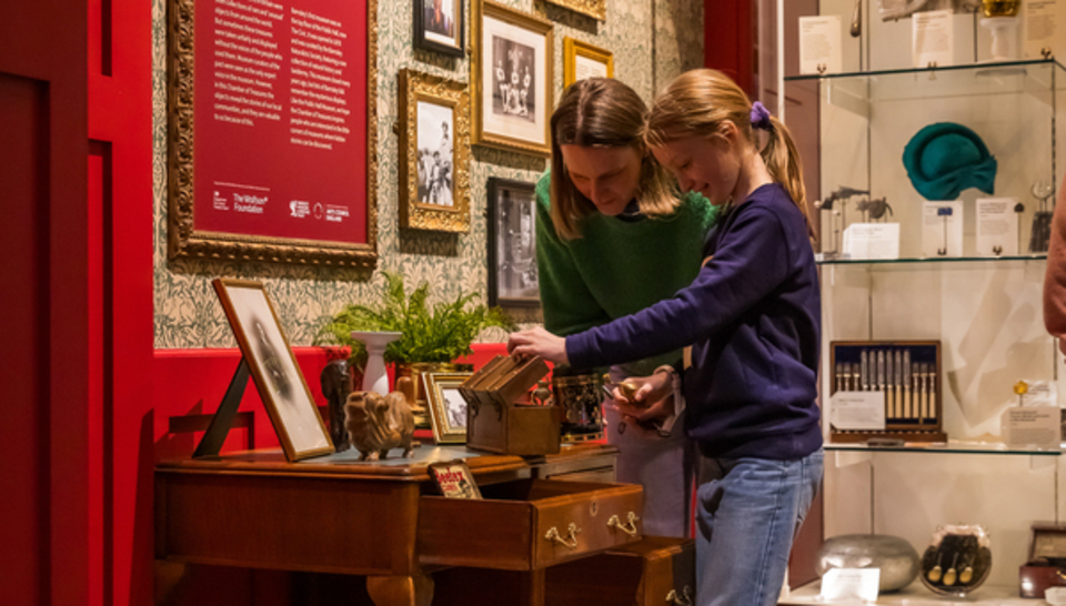 An adult and a child examine items on a small wooden desk in a museum exhibit, surrounded by framed photos, artifacts, and informational displays on the wall and shelves.